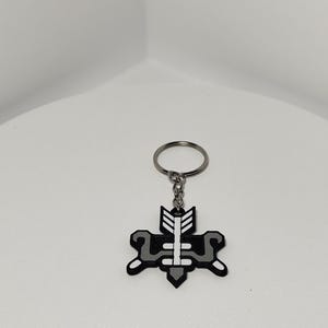 May include: A black and gray keychain with a geometric design featuring an arrow and a stylized shape. The keychain is attached to a silver metal ring. The design is centered on the keychain.