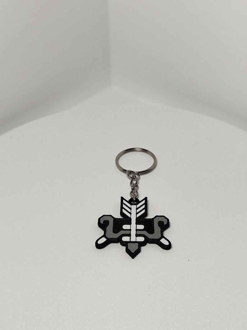 May include: A black and gray keychain with a geometric design featuring an arrow and a stylized shape. The keychain is attached to a silver metal ring. The design is centered on the keychain.