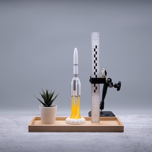 May include: A miniature rocket model with yellow flames, a small potted plant, a ruler, and a penguin figurine are arranged on a wooden tray. The rocket is white with blue and gray accents. The background is a solid gray.