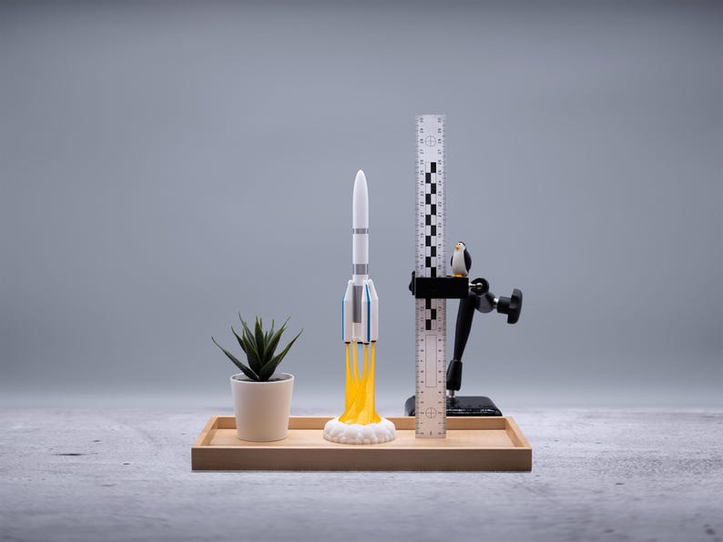 May include: A miniature rocket model with yellow flames, a small potted plant, a ruler, and a penguin figurine are arranged on a wooden tray. The rocket is white with blue and gray accents. The background is a solid gray.
