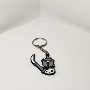 May include: A black, white, and gray keychain featuring a stylized illustration of a weapon. The keychain has a silver key ring and chain. The weapon design includes a blade and a geometric shape.