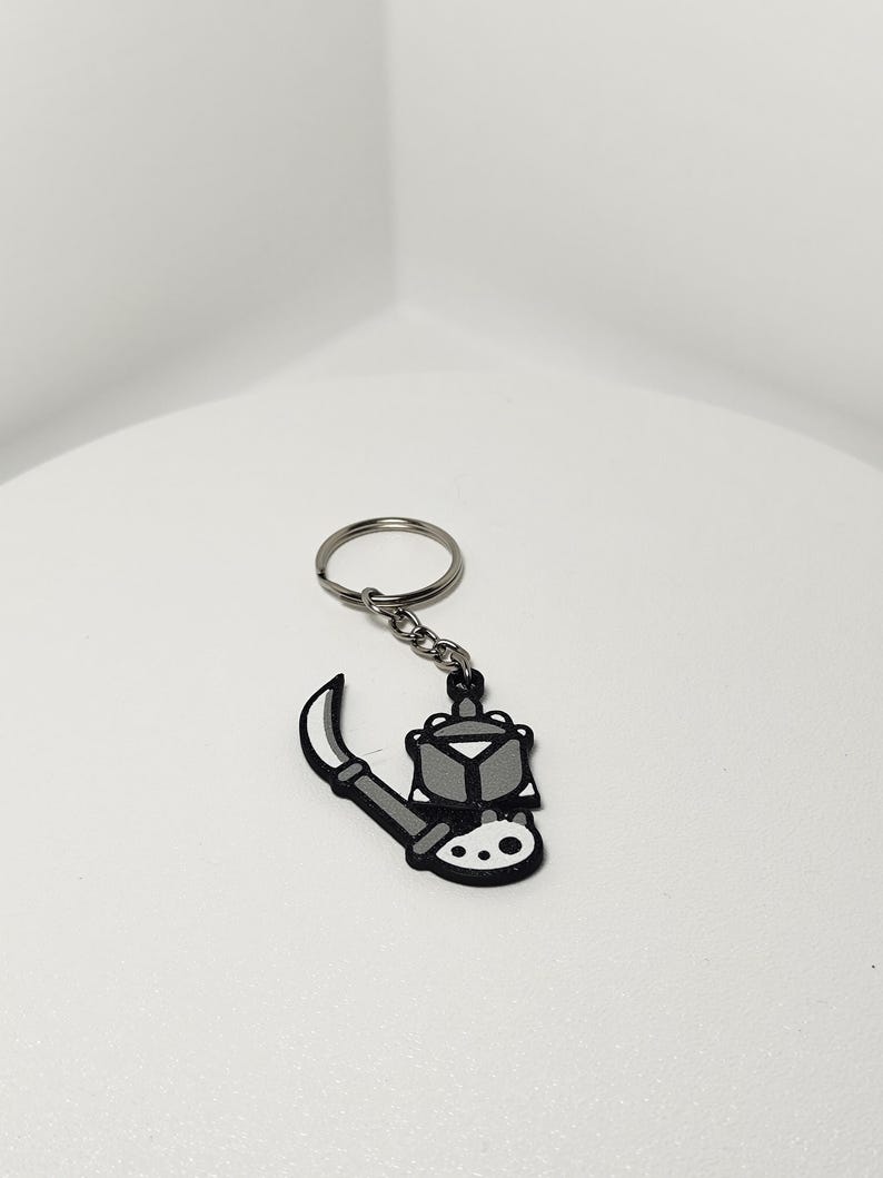 May include: A black, white, and gray keychain featuring a stylized illustration of a weapon. The keychain has a silver key ring and chain. The weapon design includes a blade and a geometric shape.
