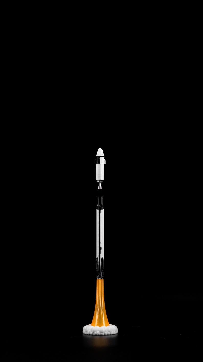 May include: A model rocket against a black background. The rocket is white and black with an orange base and a white cloud-like structure at the bottom. The rocket is standing upright.