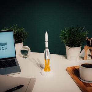 May include: A white and silver rocket model with yellow flames sits on a white desk. A laptop with the text "ANdruck" on the screen, a cup of coffee, and small potted plants are also on the desk. The background is a dark green wall.
