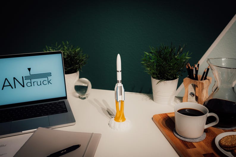 May include: A white and silver rocket model with yellow flames sits on a white desk. A laptop with the text "ANdruck" on the screen, a cup of coffee, and small potted plants are also on the desk. The background is a dark green wall.