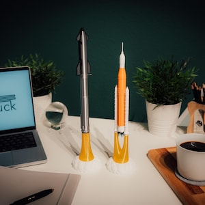 May include: Two model rockets, one silver and one orange and white, stand on a white surface. The silver rocket has a yellow base, and the orange rocket has a white base. A laptop with the text "ANdruck" on the screen is visible.