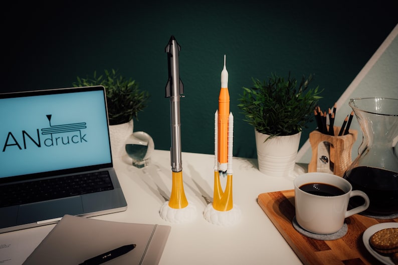 May include: Two model rockets, one silver and one orange and white, stand on a white surface. The silver rocket has a yellow base, and the orange rocket has a white base. A laptop with the text "ANdruck" on the screen is visible.