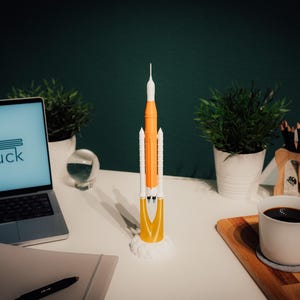 May include: A model rocket, mainly orange and white, is displayed on a white desk. The rocket is on a white base. A laptop showing "ANdruck" is also on the desk, along with plants, a coffee mug, and a notepad. The desk is in a room.