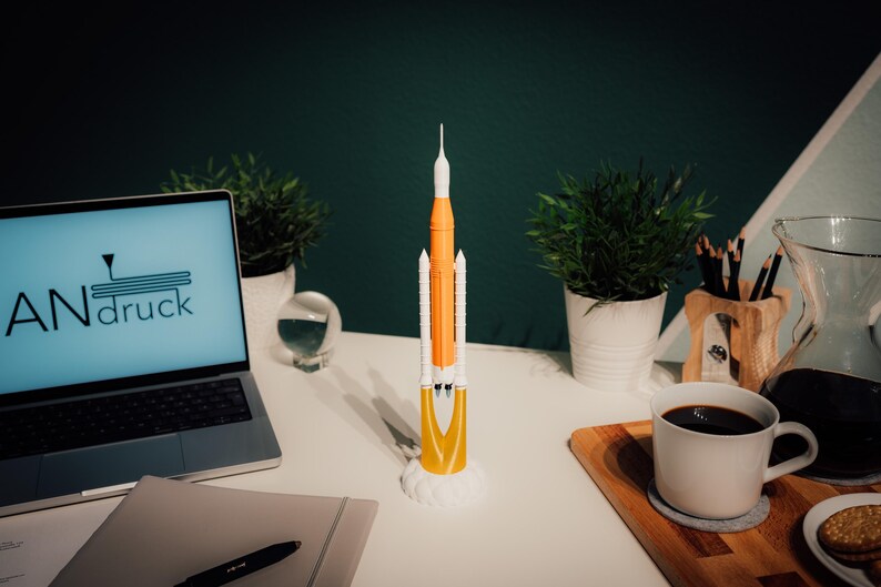 May include: A model rocket, mainly orange and white, is displayed on a white desk. The rocket is on a white base. A laptop showing "ANdruck" is also on the desk, along with plants, a coffee mug, and a notepad. The desk is in a room.