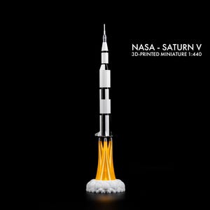 May include: A 3D-printed miniature of the NASA Saturn V rocket, scale 1:440, against a black background. The rocket is white and black with orange flames and white smoke at the base. The text "NASA - Saturn V" is displayed.