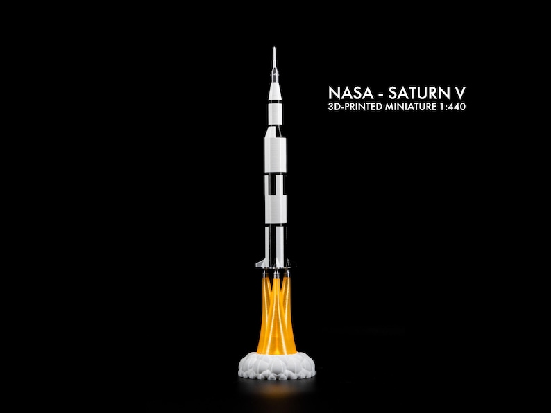 May include: A 3D-printed miniature of the NASA Saturn V rocket, scale 1:440, against a black background. The rocket is white and black with orange flames and white smoke at the base. The text "NASA - Saturn V" is displayed.