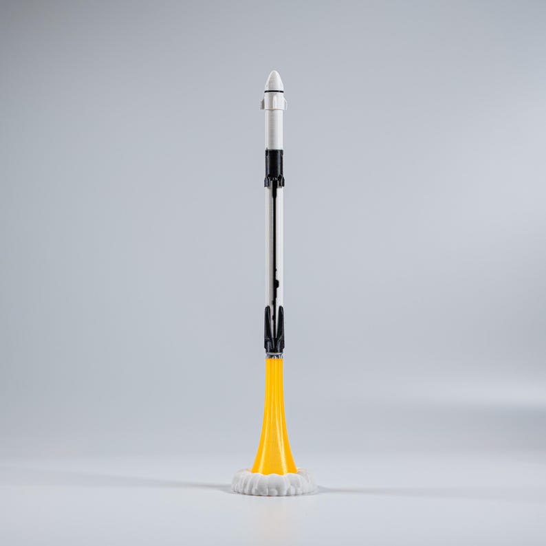 May include: A model rocket with a white nose cone, black and silver body, and yellow flames. The rocket sits on a white cloud-like base. The rocket is a miniature representation of a spacecraft.