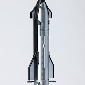 May include: A silver and black model rocket with a pointed nose and fin-like structures. The rocket has a cylindrical body with detailed paneling and a base with small, horizontal fins. The design suggests a spacecraft or aerospace theme.