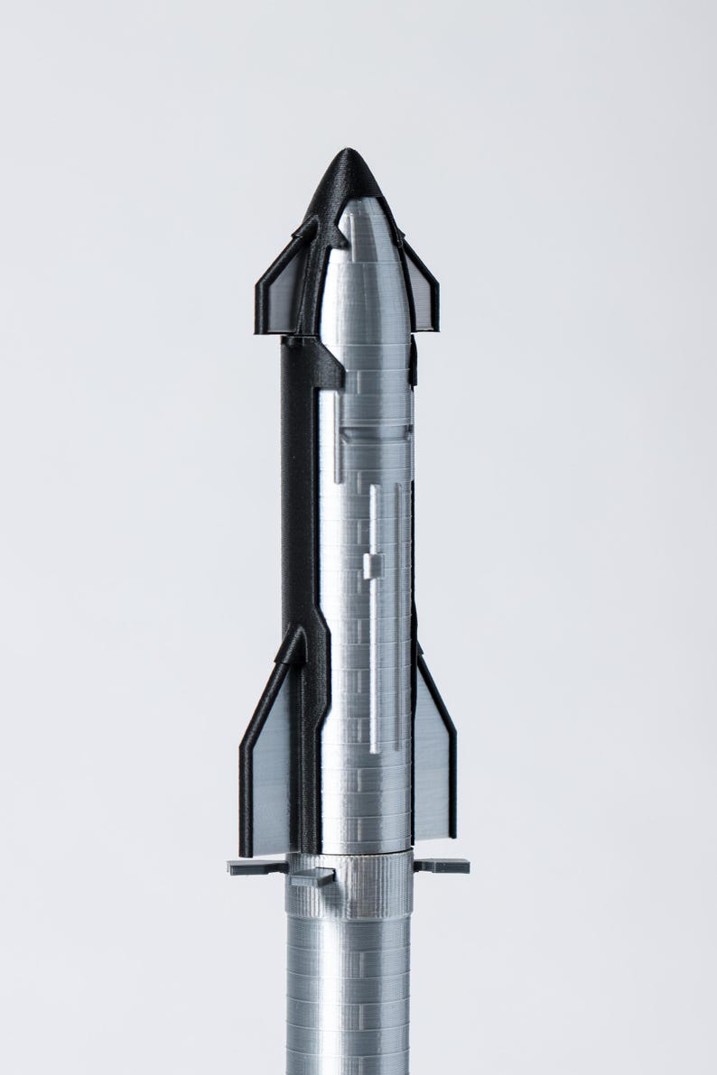 May include: A silver and black model rocket with a pointed nose and fin-like structures. The rocket has a cylindrical body with detailed paneling and a base with small, horizontal fins. The design suggests a spacecraft or aerospace theme.