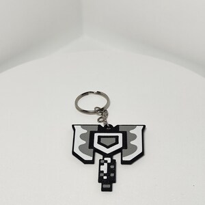 May include: A pixelated, gray and white axe-shaped keychain. The keychain features a silver key ring and a detailed design with a shield-like shape at the top and a handle at the bottom. The design is reminiscent of a video game graphic.