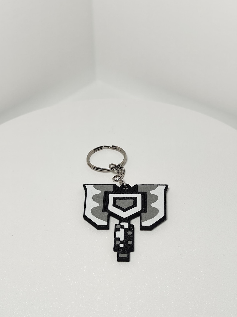 May include: A pixelated, gray and white axe-shaped keychain. The keychain features a silver key ring and a detailed design with a shield-like shape at the top and a handle at the bottom. The design is reminiscent of a video game graphic.