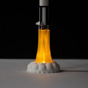May include: A decorative rocket ship lamp with a white base and a black and white rocket body. The lamp emits a bright orange glow, simulating a rocket launch. The base is white and cloud-shaped.