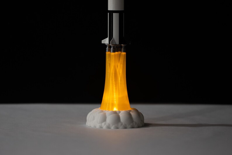 May include: A decorative rocket ship lamp with a white base and a black and white rocket body. The lamp emits a bright orange glow, simulating a rocket launch. The base is white and cloud-shaped.