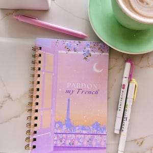 Puede incluir: Un cuaderno de espiral con una cubierta de degradado morado y naranja que presenta una ilustración de la Torre Eiffel, una luna creciente y detalles florales. El texto "PARDON my French" está impreso en el centro. Un bolígrafo rosa y dos rotuladores están cerca.