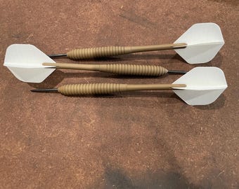 May include: Three darts with white flights and tan barrels are arranged on a brown surface. The darts have black tips and a ridged grip area. The white flights are diamond-shaped. The darts are designed for target practice.