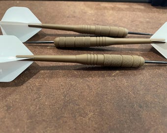 May include: Three darts with white flights and tan barrels are arranged on a brown surface. The darts have a textured grip area and a metal tip. The flights are a classic, simple design. The darts are for a game of skill.