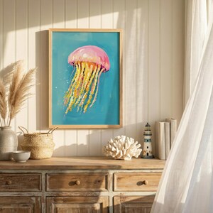 K&ouml;nnte beinhalten: Gerahmter Kunstdruck mit einem Gem&auml;lde einer Qualle. Die rosa und gelbe Qualle sticht vor einem t&uuml;rkisfarbenen Hintergrund hervor. Das Bild ist in einem hellen Holzrahmen, passend f&uuml;r eine Einrichtung im K&uuml;stenstil.