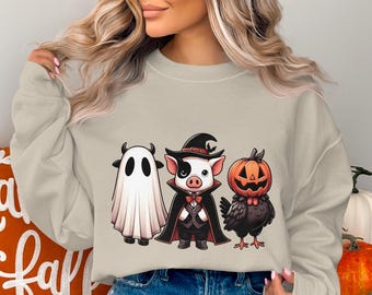Könnte beinhalten: Beiges Sweatshirt mit drei Halloween-Charakteren: einem Geist, einem Schwein mit Hexenhut und Umhang und einem Kürbis auf einem Vogel. Die Illustrationen im Cartoon-Stil sind ideal für die Herbstsaison.
