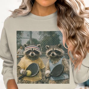 May include: Beige sweatshirt featuring a graphic of two raccoons playing paddleball. One raccoon wears a yellow shirt and headband, holding a paddle and ball. The other raccoon wears a blue tank top and holds a paddle. A chain-link fence is in the background.