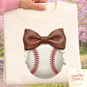 May include: Cream-coloured sweatshirt with a baseball design. A brown bow tie with a cactus and bull skull pattern sits atop a baseball with red stitching. The design includes gold paint splatters. The brand name "SUN BERRY CHARM CO" is visible.
