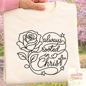 Könnte beinhalten: Cremefarbenes Sweatshirt mit schwarzem Blumendesign und dem Schriftzug "always rooted in Christ". Das Design umfasst eine Rose, Blätter, Sterne und ein Kreuz. Das Sweatshirt wird von einer Person gehalten.