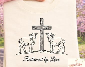 May include: Cream-coloured sweatshirt with a black line drawing of a wooden cross and two lambs. The text "Redeemed by Love" is below the drawing. The sweatshirt is held by a person.