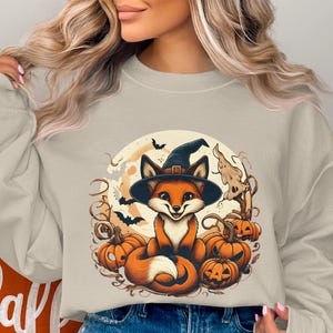 Puede incluir: Sudadera beige con un diseño de Halloween. El gráfico muestra un zorro de dibujos animados con un sombrero de bruja, rodeado de calabazas, murciélagos y una luna llena. El zorro es naranja y blanco, y el sombrero es negro. El diseño es caprichoso.