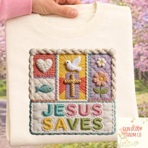 May include: Cream sweatshirt with a colourful embroidered design. The design features a heart, dove, cross, fish, and flower, with the words "JESUS SAVES" in a rainbow of colours. The sweatshirt is held by a person.