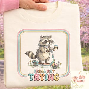 Puede incluir: Sudadera color crema con un mapache montado en un scooter y sosteniendo una taza de café. El diseño incluye el texto "FERAL BUT TRYING" en letras coloridas, enmarcado por rayas pastel. La sudadera tiene cuello redondo.