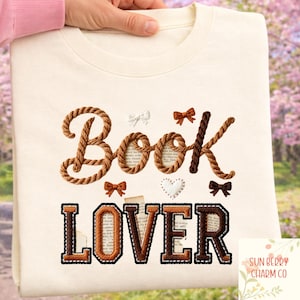Peut inclure: Sweat-shirt crème avec les mots "Book Lover" brodés en marron et noir. Le mot "Book" est écrit avec une police en forme de corde, et le mot "Lover" est en police de style universitaire. Petits nœuds décoratifs.