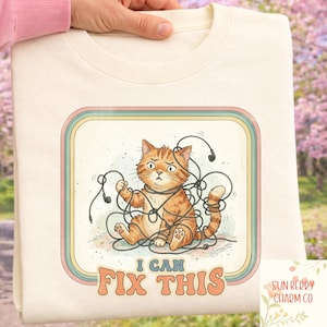 May include: Cream-colored sweatshirt featuring a distressed orange tabby cat tangled in black headphone cords. The cat has a worried expression. Text reads "I CAN FIX THIS" in a retro font. The design is framed in a colorful border.