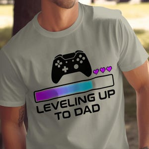 May include: A light grey t-shirt with a black video game controller graphic above a purple and blue progress bar. The text "LEVELING UP TO DAD" is below the bar.