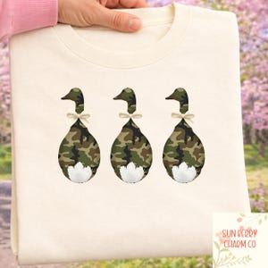 May include: Cream-colored sweatshirt featuring three duck designs. Each duck has a camouflage body, a white fluffy tail, and a bow tie. The ducks are facing away from the viewer. The sweatshirt is held by a person.