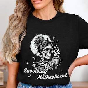 May include: Black t-shirt featuring a skeleton with a bandana and a coffee cup, with the words "Surviving Motherhood" in white. The skeleton is set against a moon and star background. A humorous graphic tee.