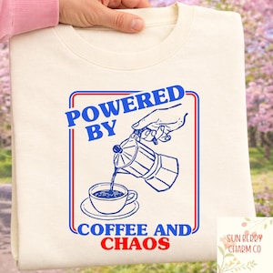 以下が含まれることがあります： クリーム色のスウェットシャツ。青と赤のグラフィックで、「POWERED BY COFFEE AND CHAOS」と書かれ、コーヒーがカップに注がれるイラストが描かれています。ブランドロゴは右下にあります。