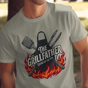 Peut inclure: T-shirt gris clair avec un motif sur le thème du barbecue. Le motif comprend le texte "The Grillfather" au-dessus de "Legend of the Pit", un tablier, une spatule, des pinces et des flammes.