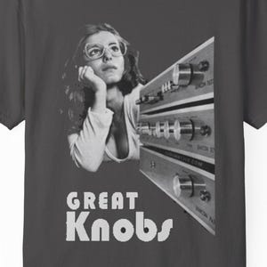 May include: Dark gray t-shirt featuring a black and white graphic. The graphic includes a woman with glasses and a vintage stereo component. The text "GREAT Knobs" is printed below the image.