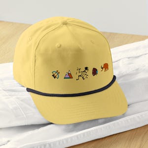 Puede incluir: Gorra de béisbol amarilla mostaza con ribete de cuerda negra. La gorra presenta una serie de gráficos coloridos de inspiración nativa americana, que incluyen un arco y flecha, montañas, una figura corriendo, un tocado y un elefante.
