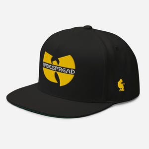 May include: A black snapback cap featuring a yellow embroidered logo on the front with the word WIDESPREAD in white text across the center. A small yellow embroidered figure sitting on a stool is visible on the side of the cap.