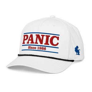 May include: A white baseball cap featuring a black rope detail across the brim. The front displays the red embroidered text PANIC above the red text Since 1986, framed by blue horizontal lines. A small blue silhouette graphic is on the side.
