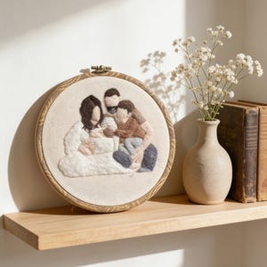 May include: A circular, embroidered family portrait in a wooden hoop. The artwork shows a family of four in neutral tones. The hoop is displayed on a wooden shelf, next to a vase of white flowers and two antique books.