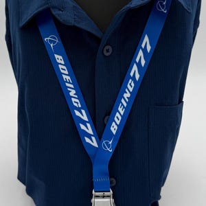 May include: A blue lanyard with the text "BOEING 777" in white. The lanyard has a silver-colored metal clasp and a black cord with a key ring. The lanyard is displayed on a blue button-down shirt.