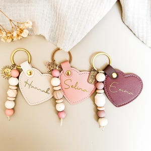 Könnte beinhalten: Drei herzförmige Schlüsselanhänger in Hellrosa, Rosa und Lila. Jeder Schlüsselanhänger hat einen goldenen Ring, einen Perlenquasten und einen Namen in goldener Schrift: Hannah, Selina und Emma. Ein Sonnenanhänger hängt am ersten Schlüsselanhänger.