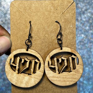 May include: A pair of wooden earrings with a circular design. Each earring features the number "420" cut out in a stylized font. The earrings are attached to black metal hooks and are displayed on a brown cardboard backing.