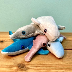 May include: A pile of plush toy whales in various colors: blue, light blue, gray, pink, and white. Each whale has a small, round, black eye and a small fin. The toys are on a wooden surface.